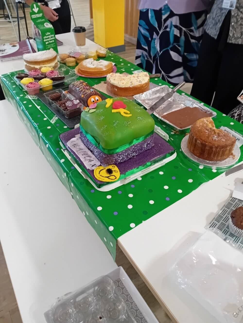 A table with various cakes and pastries, including a green frosted cake with a cartoon figure on top amidst other baked goods. - Home Instead Bournemouth & Christchurch
