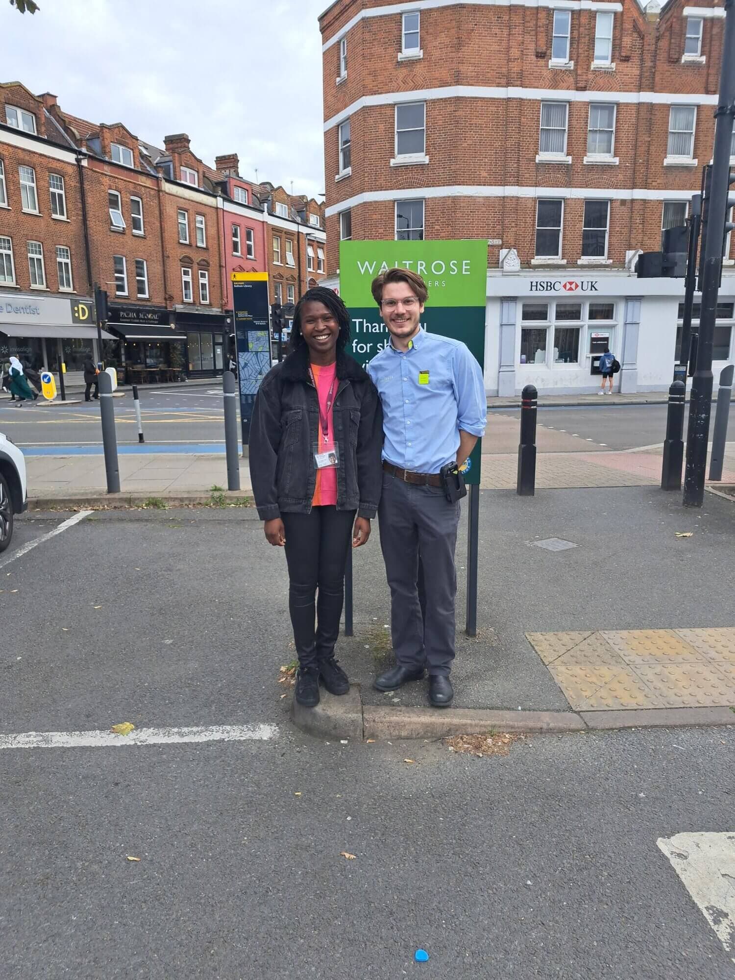 Two people standing in a parking lot next to a sign that reads "Waitrose" with buildings and shops in the background. - Home Instead