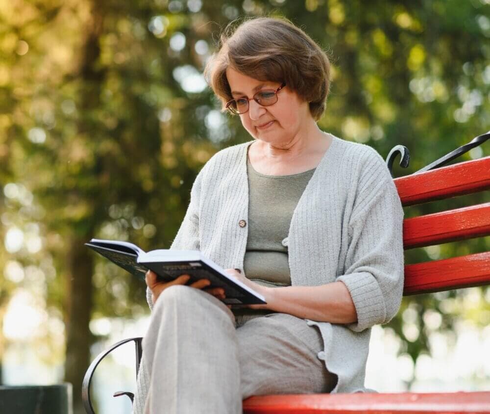 Elderly woman with short hair and glasses reading a book while sitting on a red park bench in a green, outdoor setting. - Home Instead