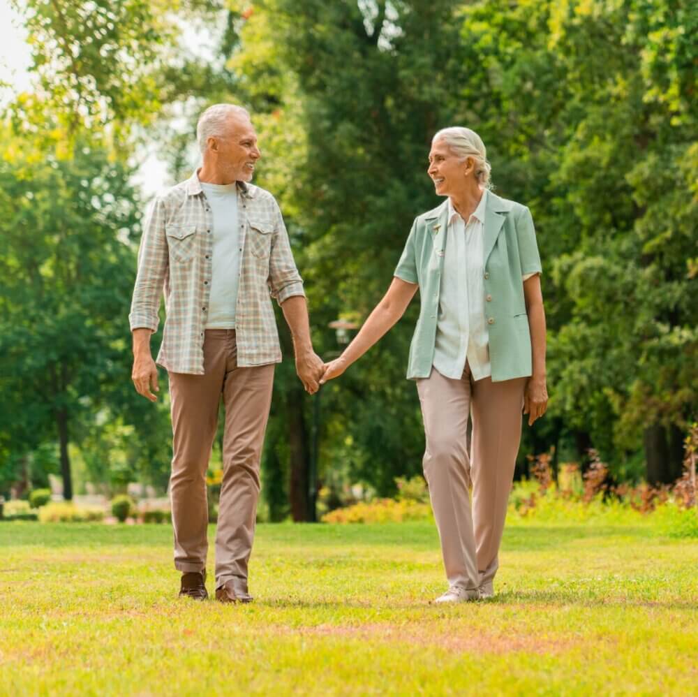 Older couple walking hand-in-hand in a sunny park, surrounded by green trees and grass. - Home Instead