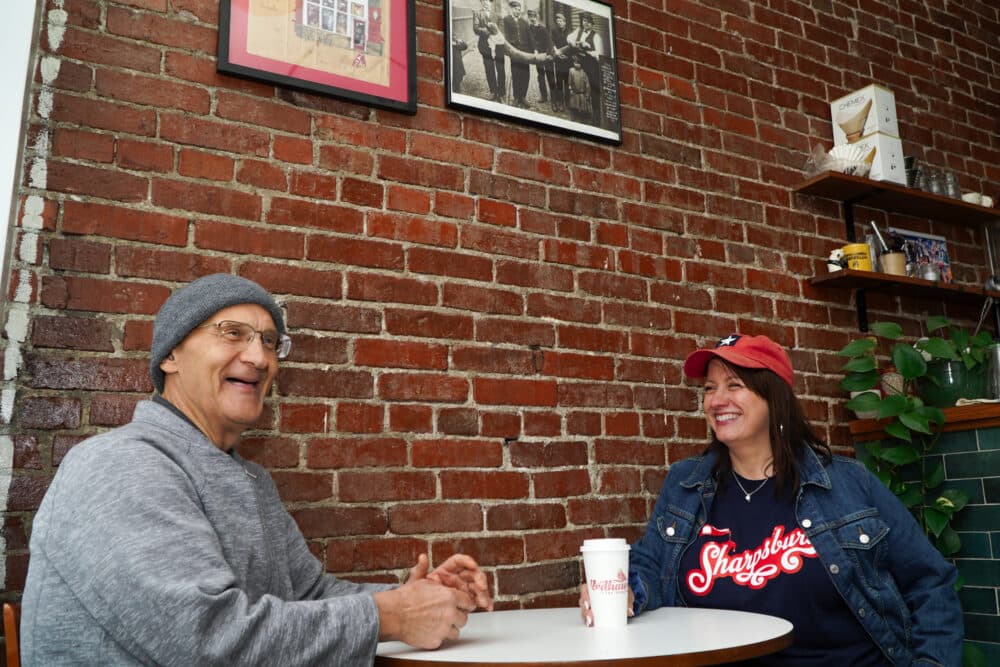 Two people smile and chat at a small table in a cozy café with brick walls and framed photos. - Home Instead