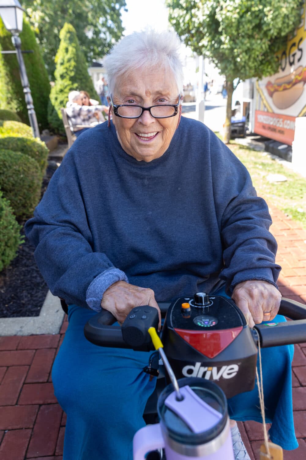 Smiling older woman with glasses sits on a mobility scooter outdoors near bushes and a hot dog stand. - Home Instead