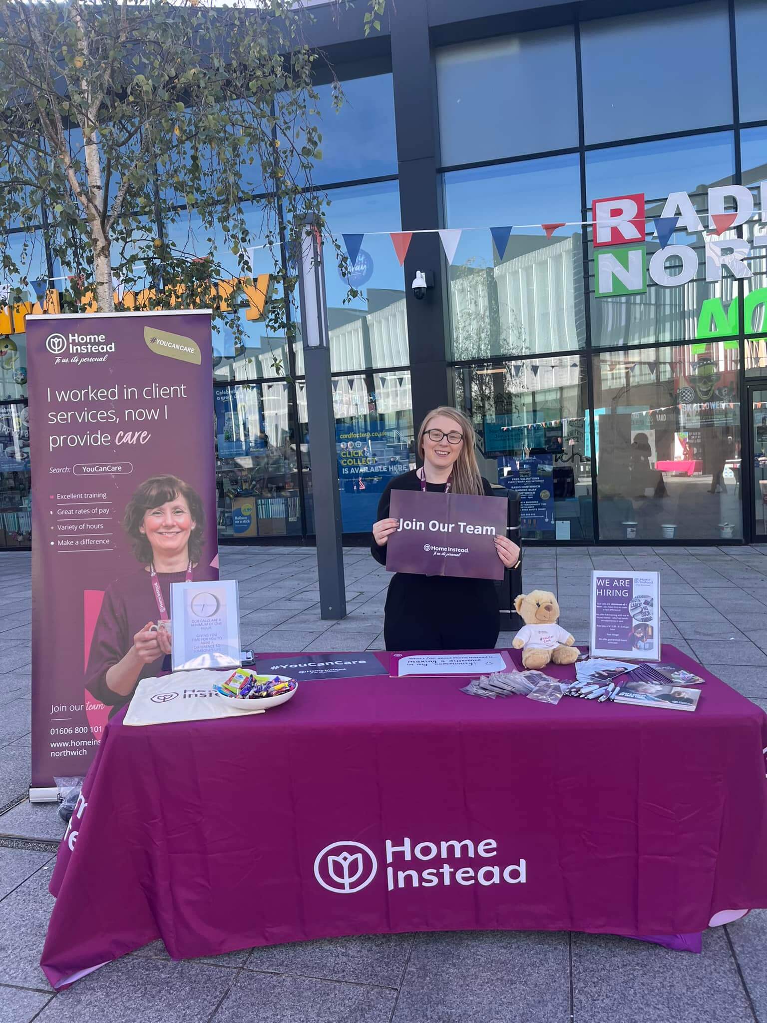 A woman stands behind a table displaying Home Instead materials, holding a "Join Our Team" sign in front of an office building. - Home Instead