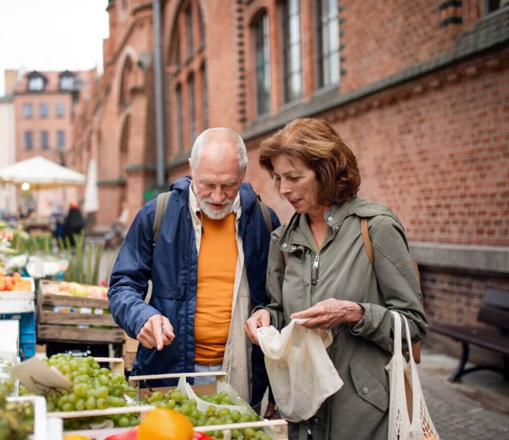An older couple shops for produce at an outdoor market stall with brick buildings in the background. - Home Instead