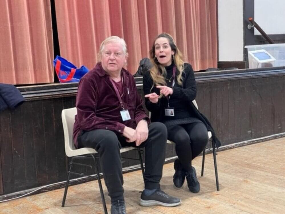 Two people sitting on chairs in a hall, with one gesturing while speaking, and the other looking ahead. - Home Instead