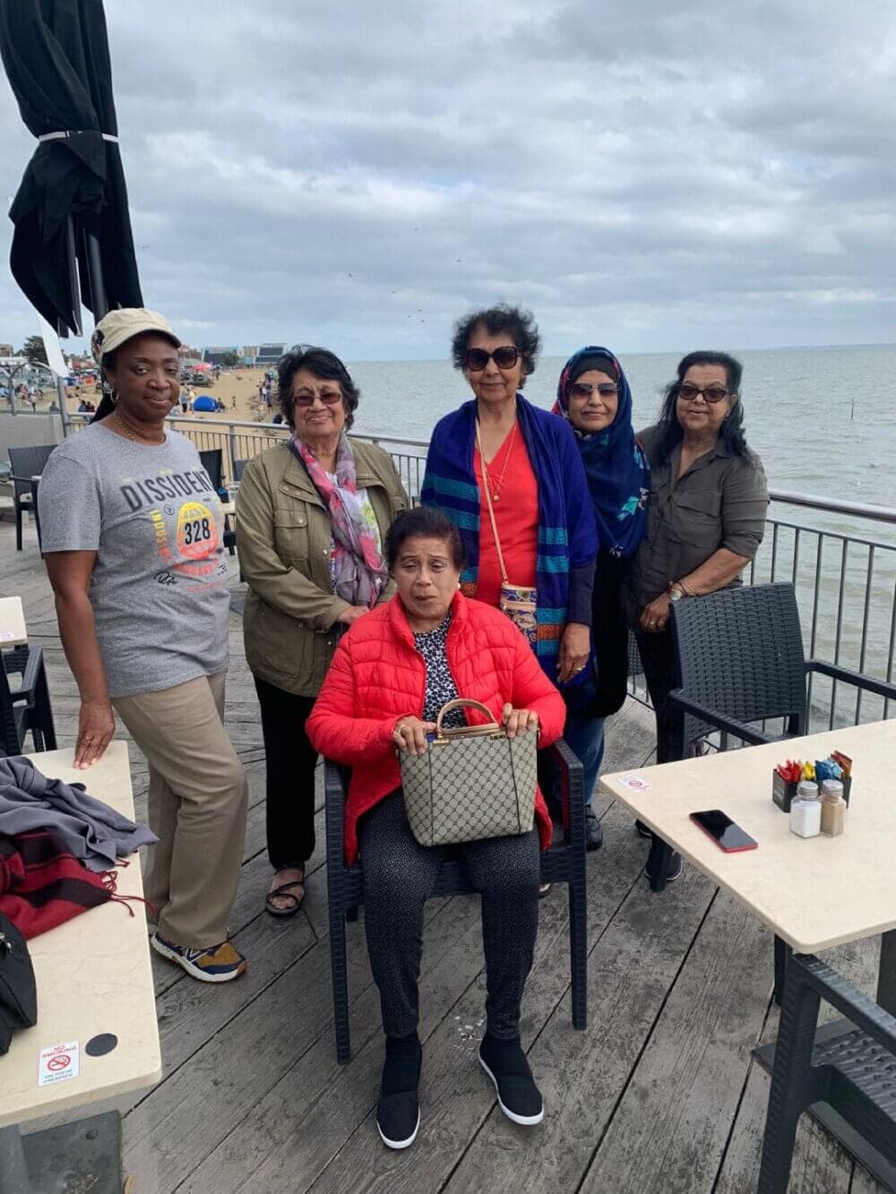 Six women are gathered at an outdoor seaside restaurant, with one seated and five standing behind her, all smiling. - Home Instead