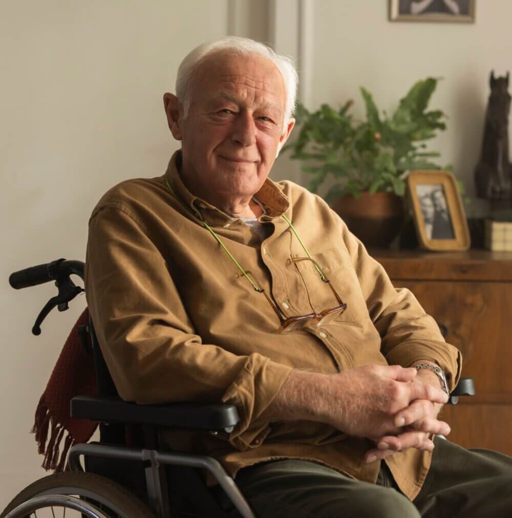 Elderly man with white hair sits smiling in a wheelchair, wearing a brown shirt with glasses hanging around his neck. - Home Instead