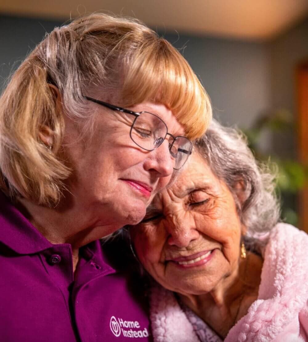 Two elderly women sharing a heartfelt hug, one wearing a "Home Instead" shirt. - Home Instead