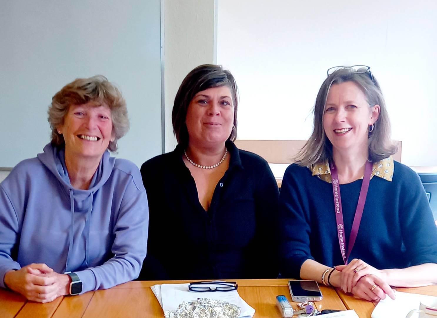Three women sitting at a table, smiling. A plate of food, a phone, and glasses are on the table in front of them. - Home Instead
