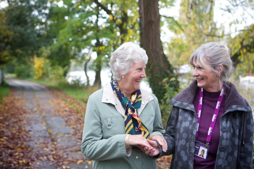 Two elderly women walk and chat on a leaf-strewn path surrounded by trees near a body of water. - Home Instead