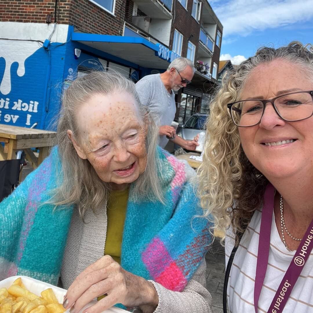 Two women smiling at an outdoor table with food; an older woman holding fries and a younger woman wearing glasses. - Home Instead
