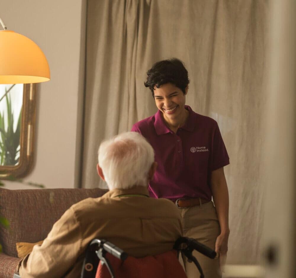 A caregiver smiles while interacting with an elderly person in a wheelchair in a warmly lit room. - Home Instead