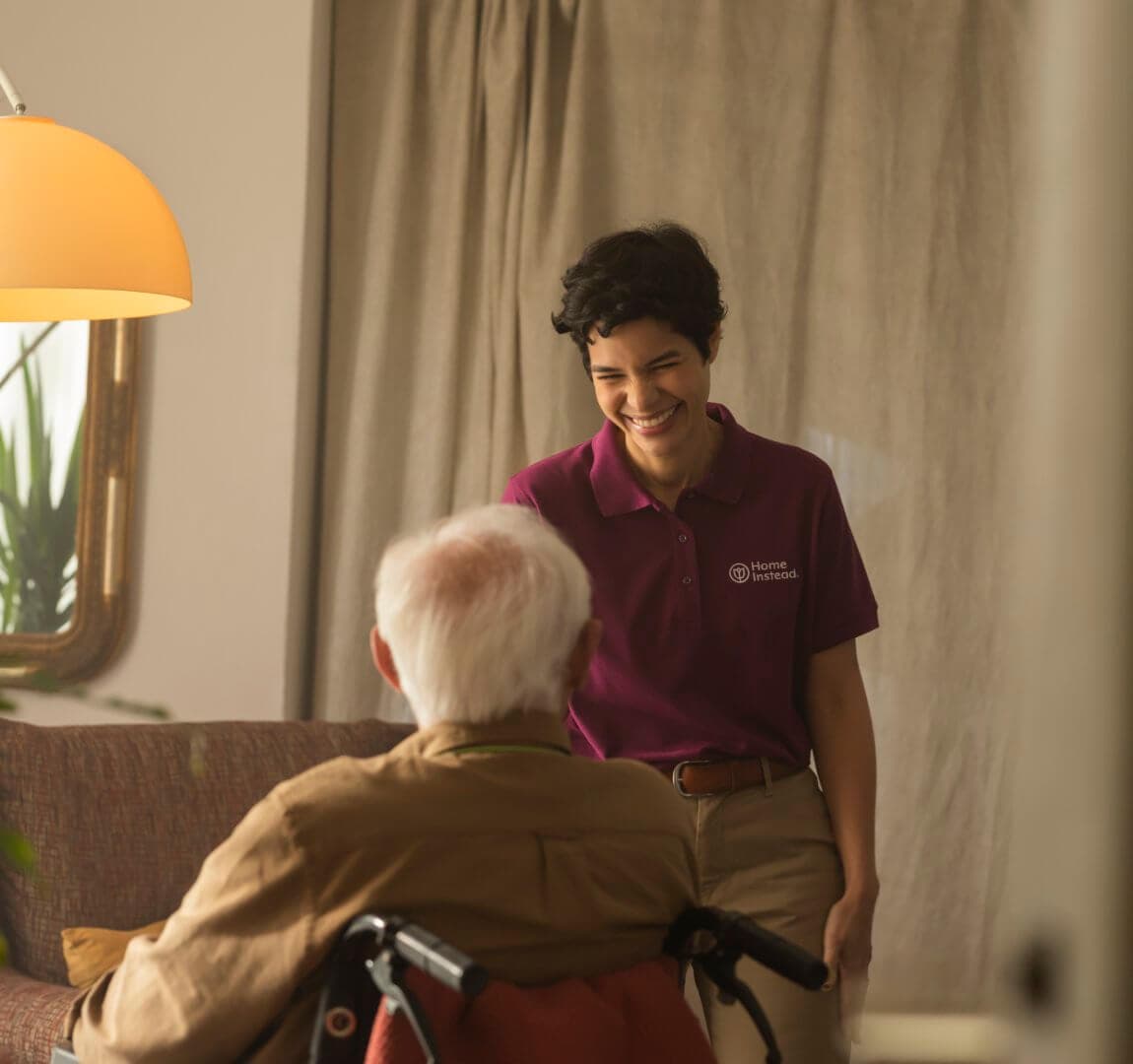 A caregiver smiles while interacting with an elderly person in a wheelchair in a warmly lit room. - Home Instead