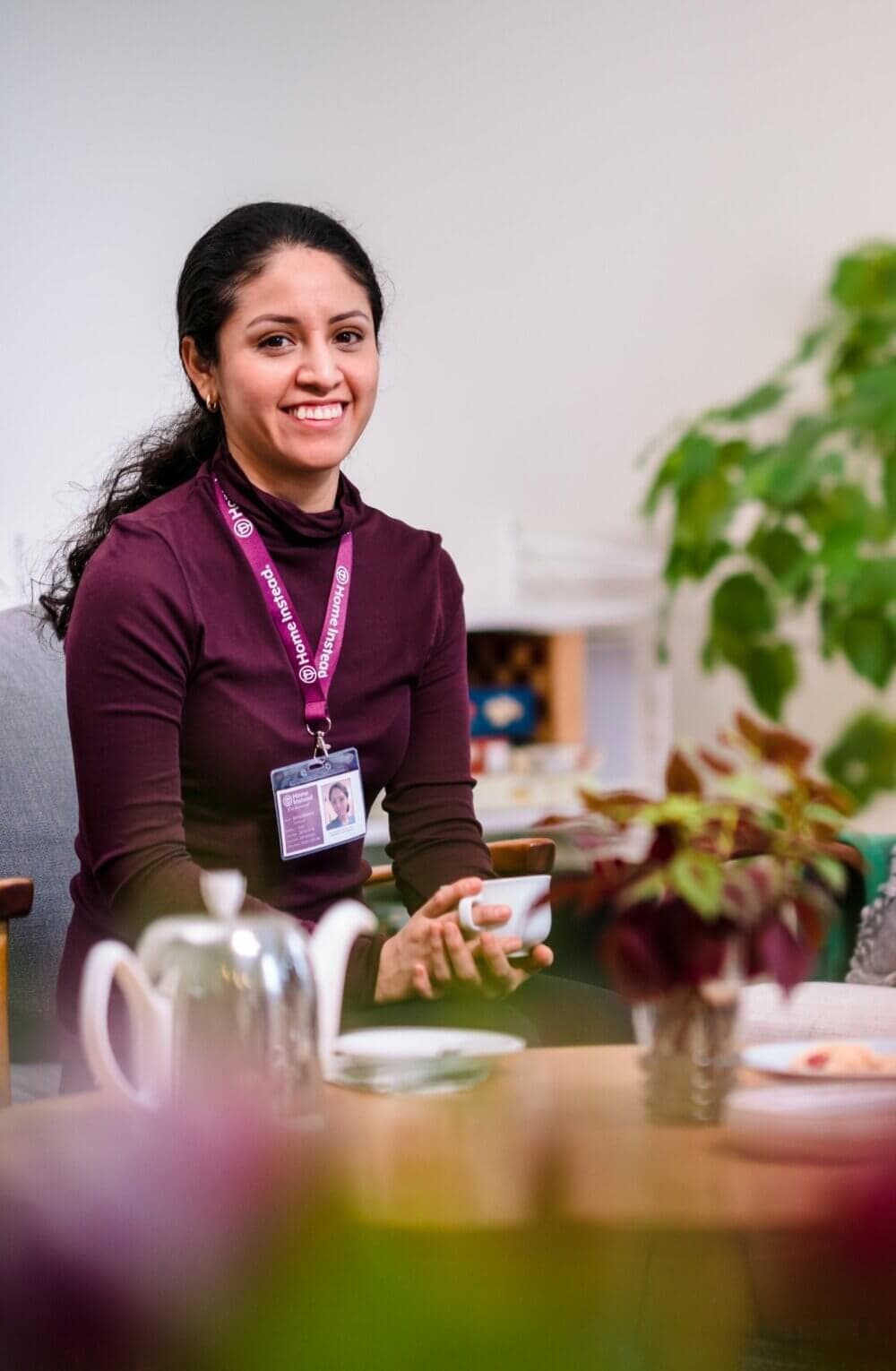 A smiling woman wearing a name badge and lanyard sits at a table with a coffee cup, surrounded by greenery. - Home Instead