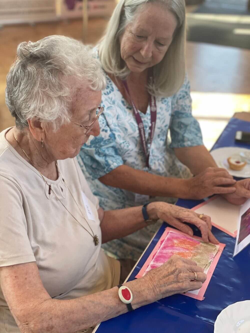Two elderly women engaged in an art project, one helping the other with a colorful piece of paper. - Home Instead