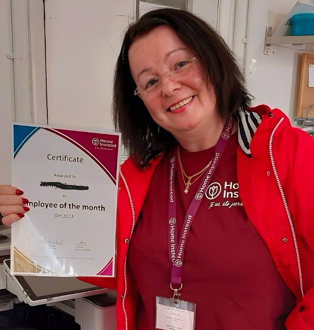 A smiling woman holds an "Employee of the Month" certificate while wearing a red jacket and lanyard in an office. - Home Instead