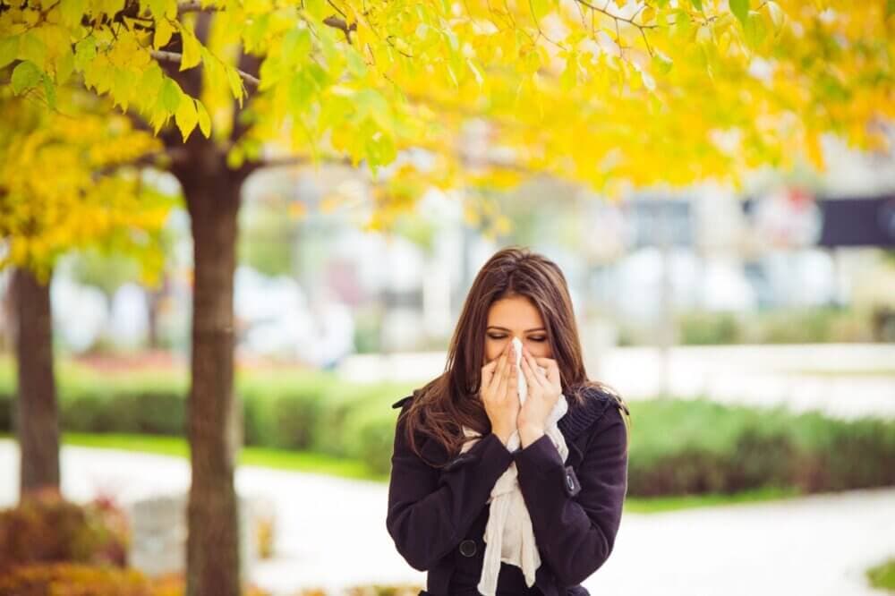Woman in a park sneezing into a tissue under autumn foliage, wearing a coat and scarf. - Home Instead