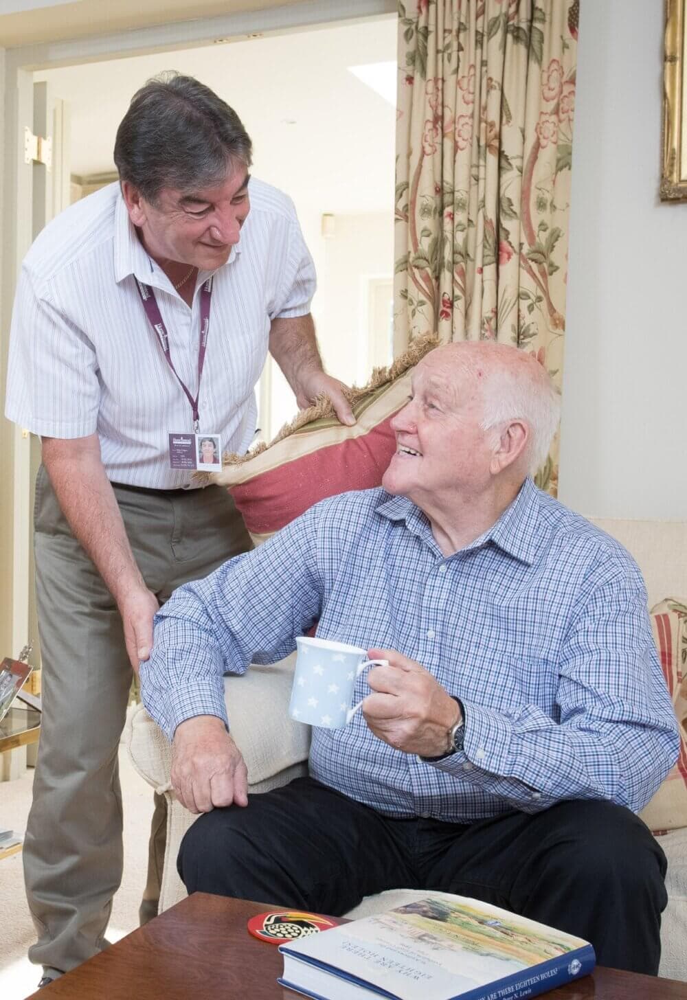 Elderly man with a mug, sitting and smiling at a standing man in uniform badge, inside a cozy room. - Home Instead