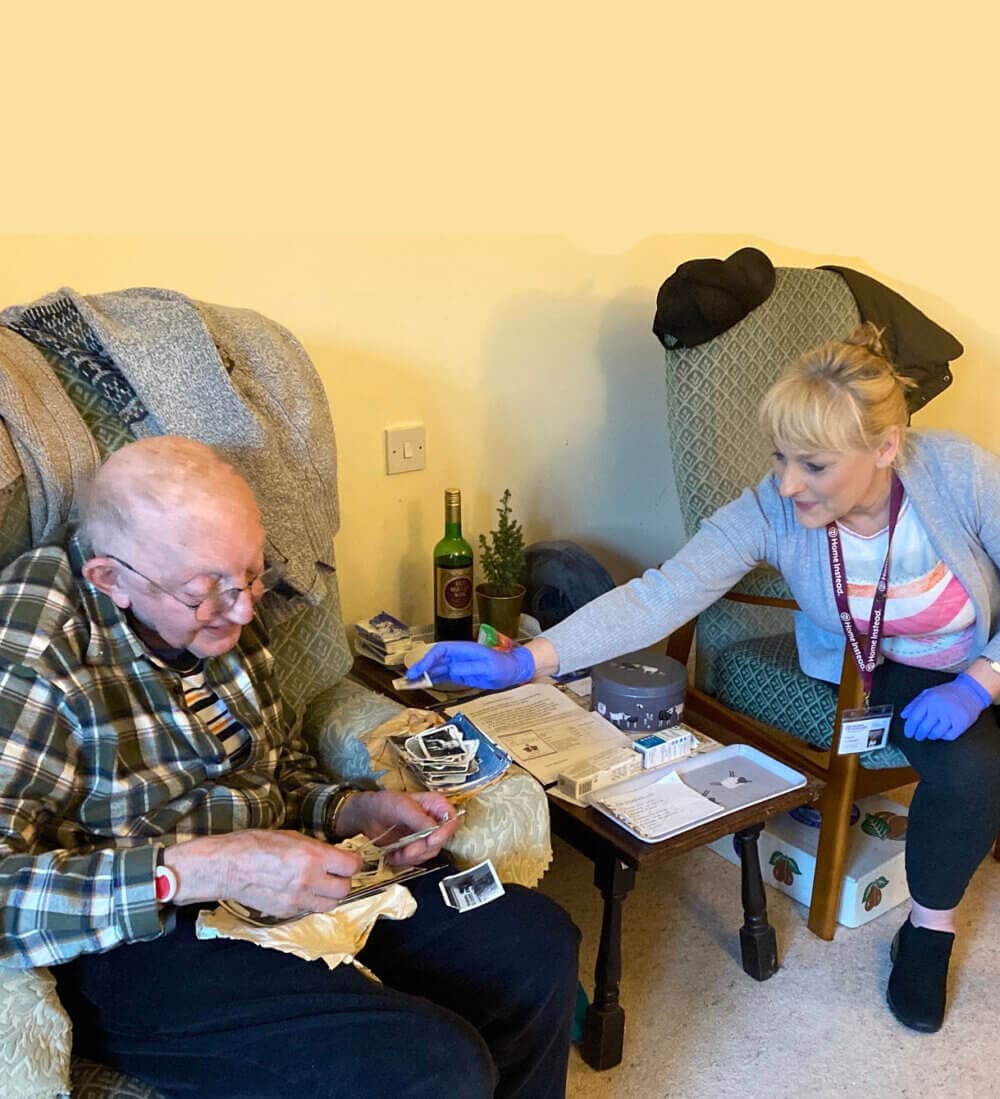 A senior man looks at photos while a caregiver, seated beside him, reaches over with a warm smile. - Home Instead