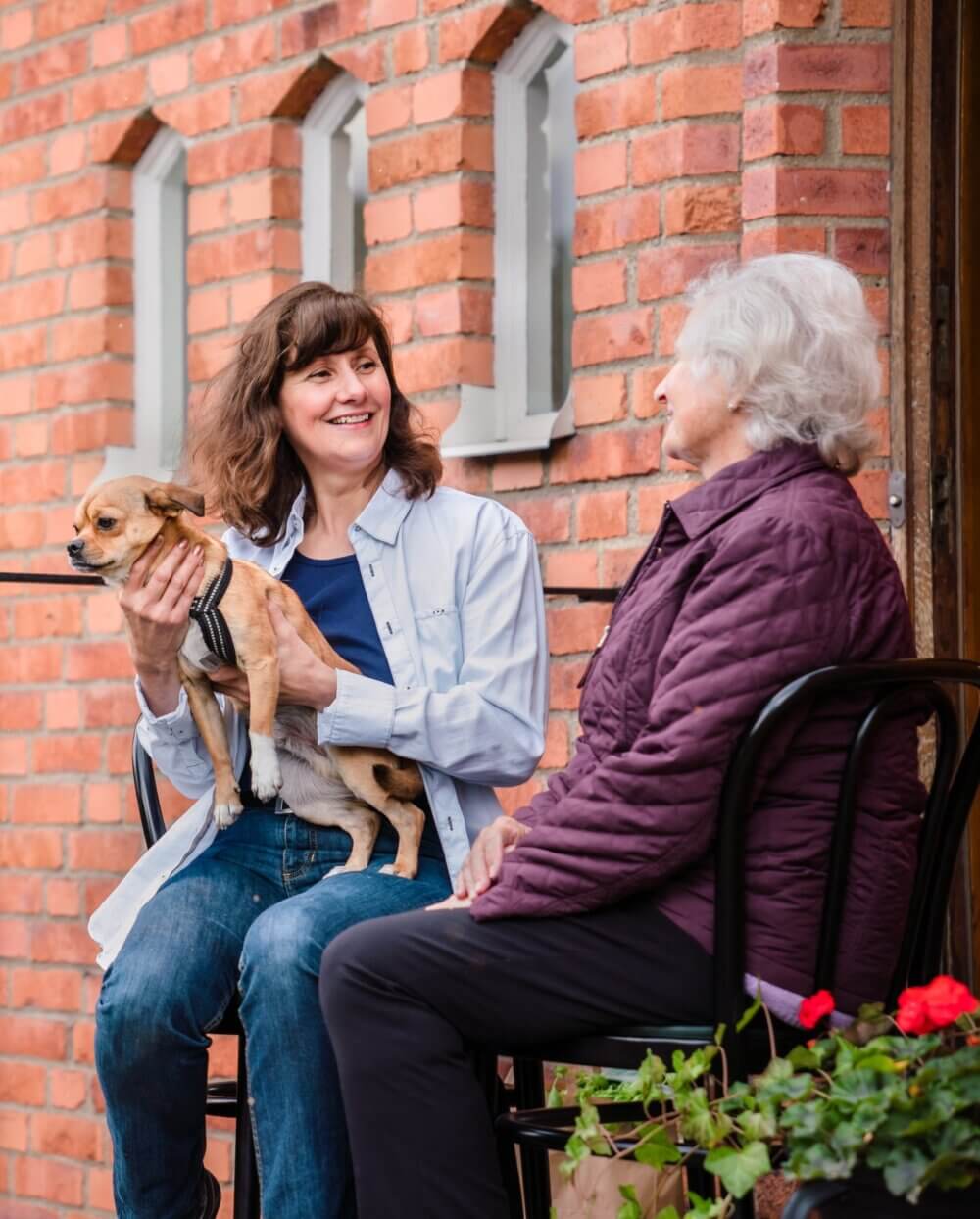 Two women sitting outside, one holding a small dog, with a brick wall and windows in the background. - Home Instead