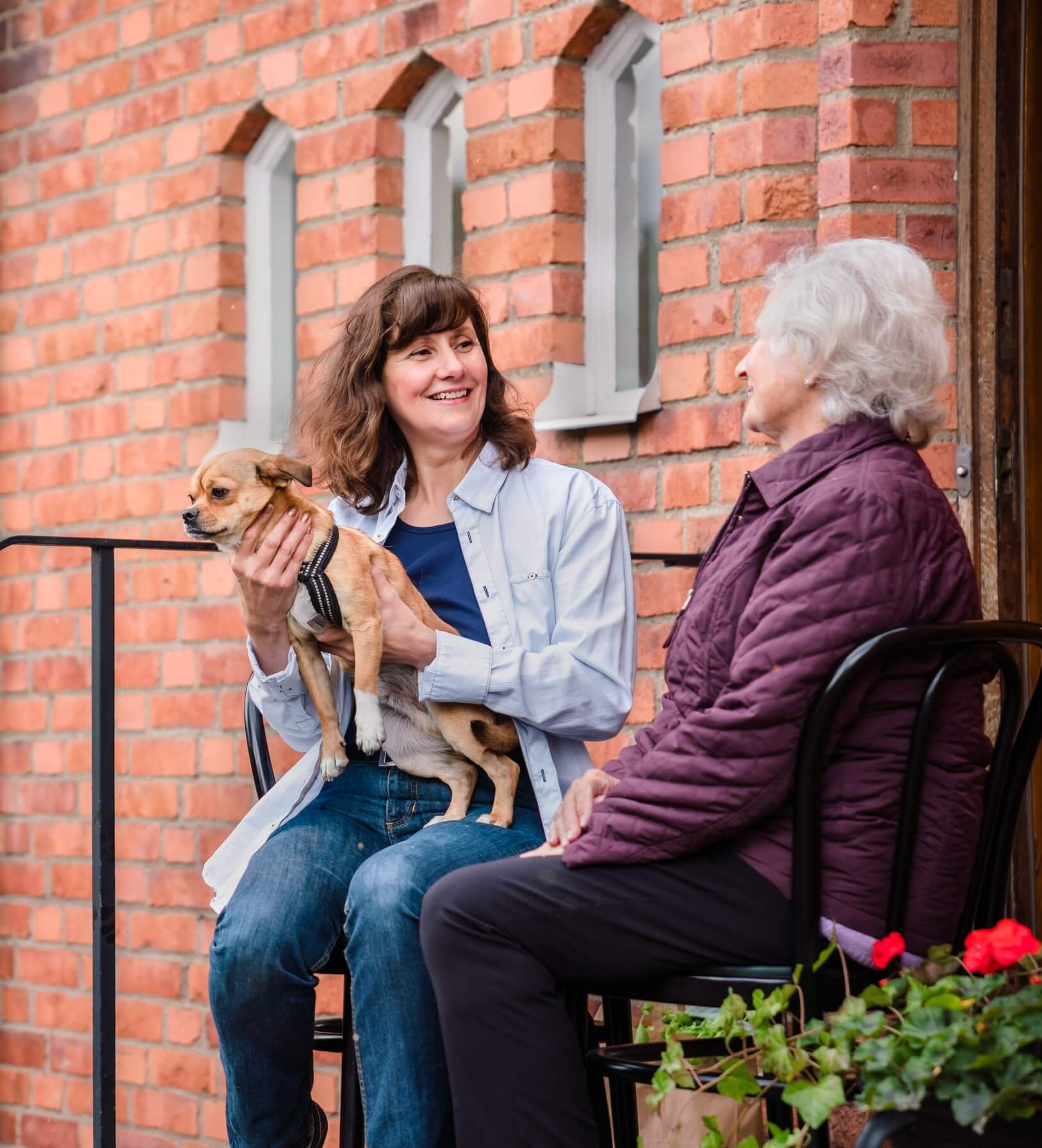 Two women, one holding a small dog, sit and talk on a porch with a brick wall background and flowers in the foreground. - Home Instead
