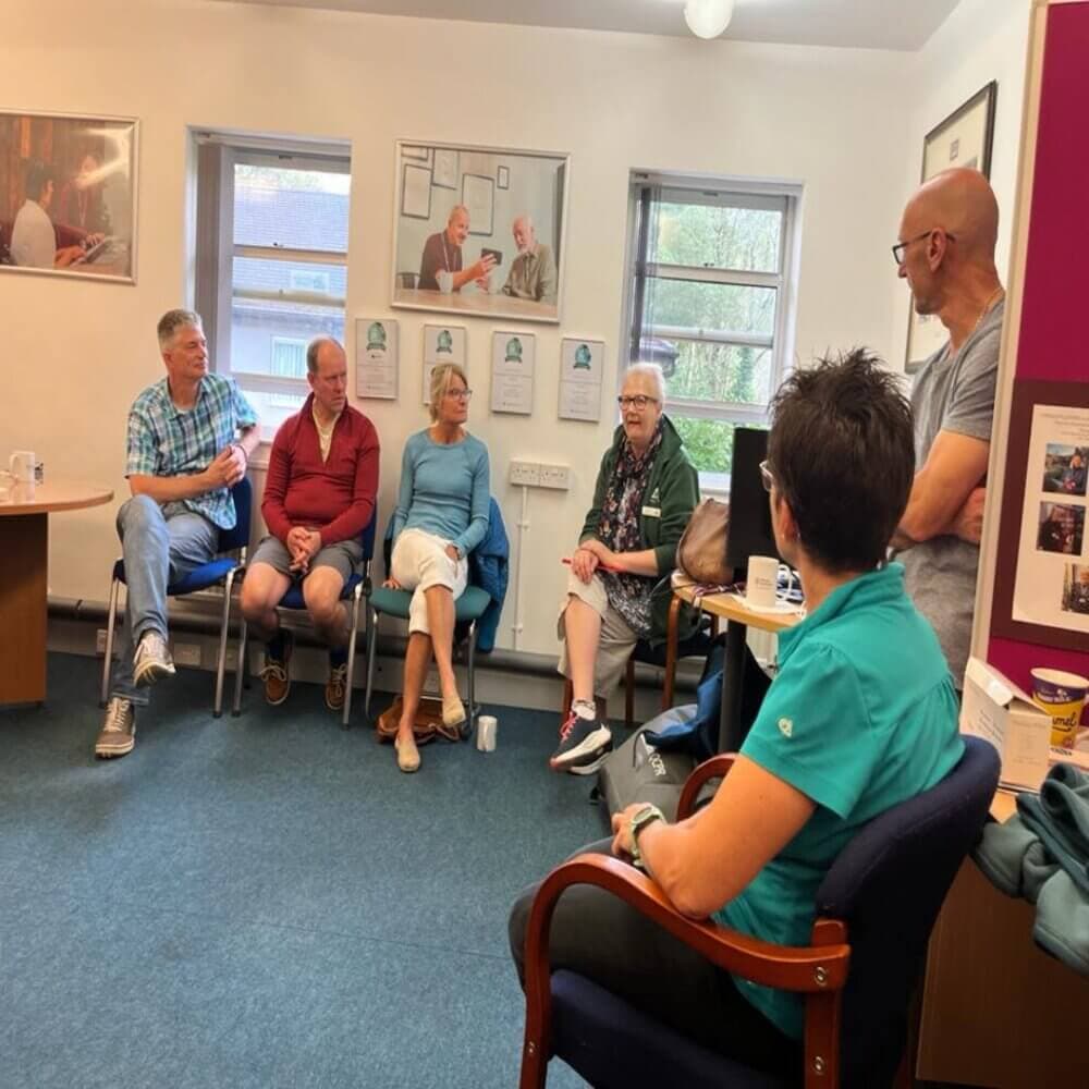 A group of people sit in a circle talking in a room with windows and framed photos on the wall. - Home Instead