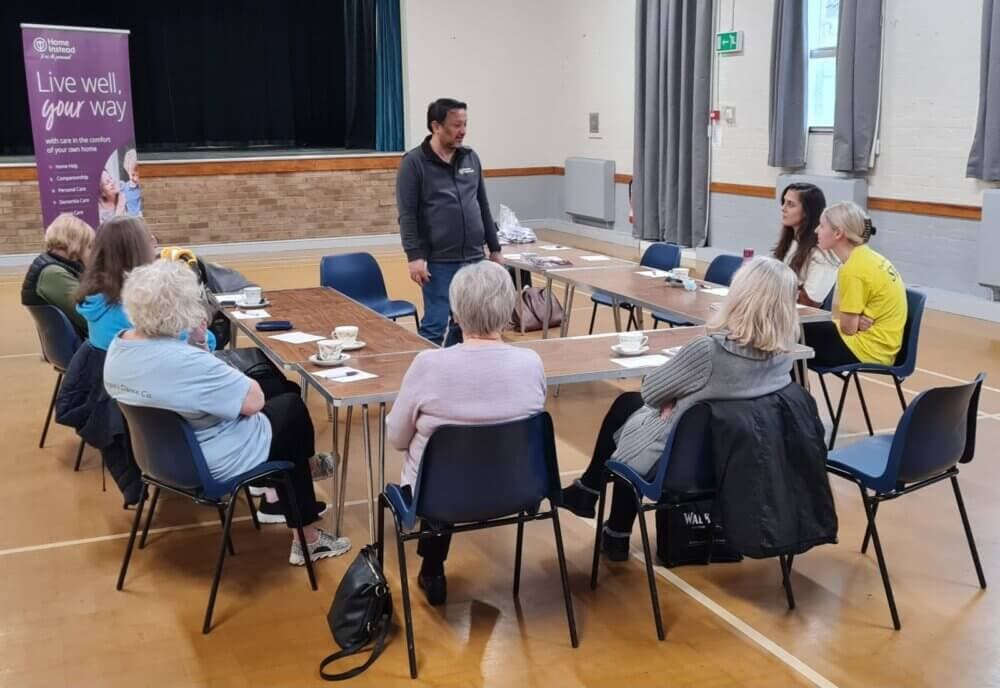 A man speaks to a group of seated people in a community hall, with a banner in the background that says "Live well, your way. - Home Instead