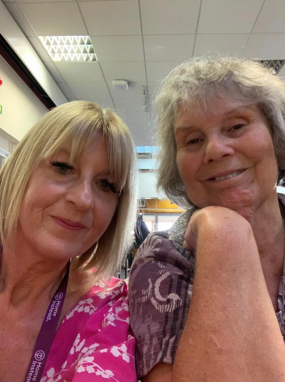 Two women sitting indoors taking a selfie, both smiling. One has short blonde hair and the other has short gray hair. - Home Instead