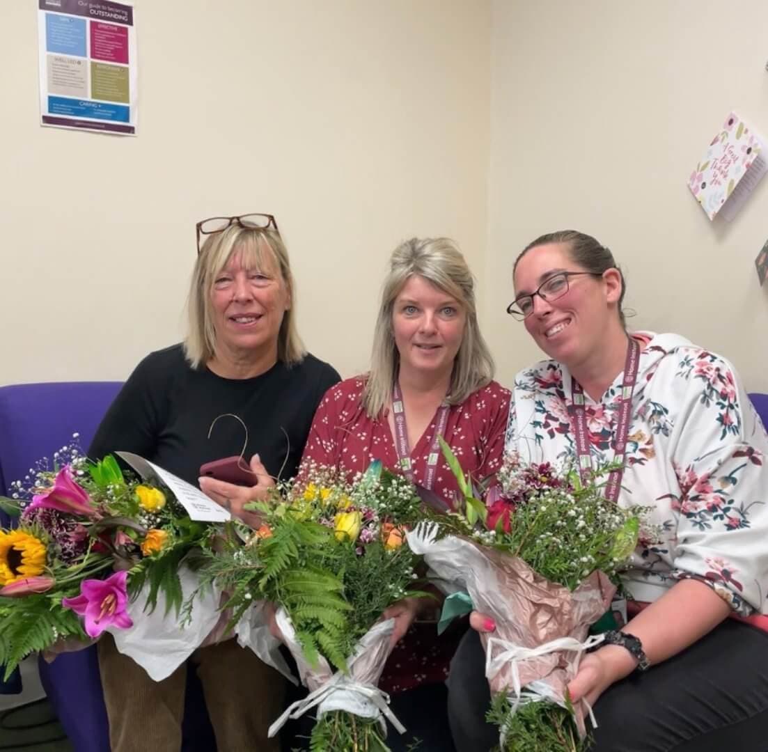 Three women smiling and holding bouquets of flowers while sitting on a purple couch in an office setting. - Home Instead
