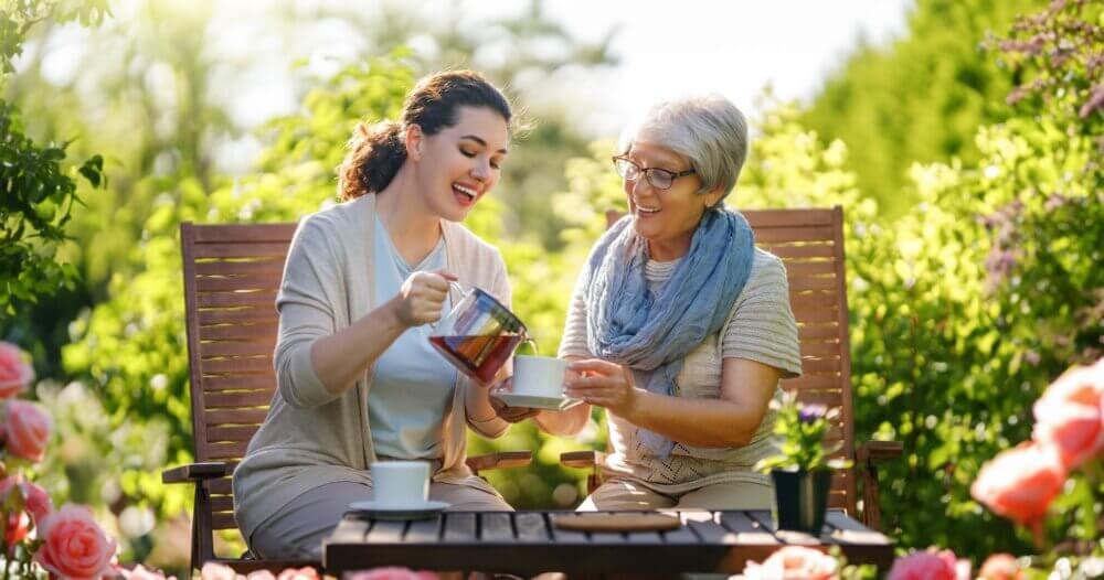 Two women enjoying tea outside, sitting on a bench in a blooming garden, surrounded by vibrant flowers. - Home Instead