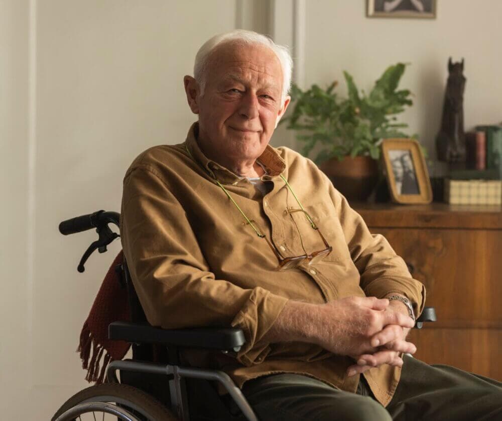 Elderly man with white hair sitting in a wheelchair, wearing a brown shirt and glasses, with plants and photos in the background. - Home Instead