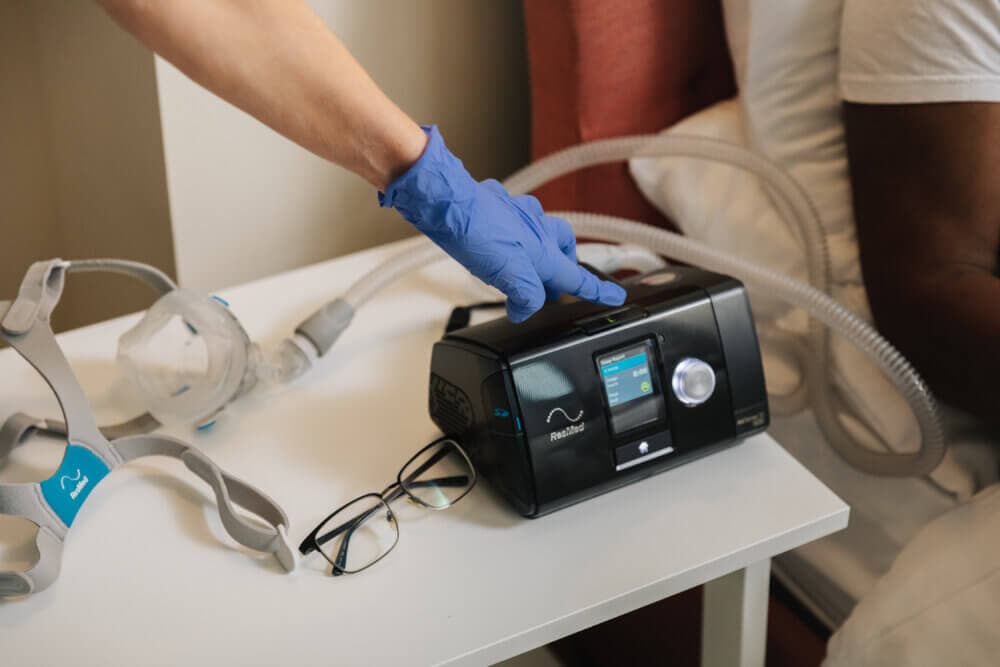 A hand in a blue glove operates a CPAP machine beside a mask and glasses on a bedside table. - Home Instead