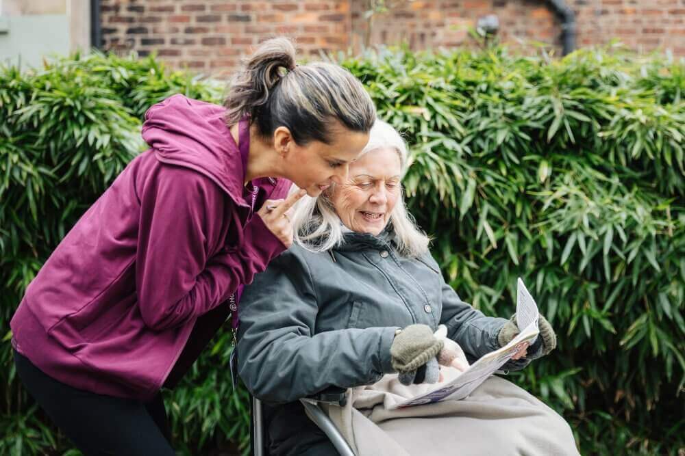 A woman leans over to chat with an elderly woman in a wheelchair who is holding a newspaper, both smiling. - Home Instead