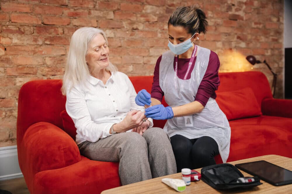 Healthcare worker in PPE giving medication to an elderly woman on a red couch with a brick wall background. - Home Instead