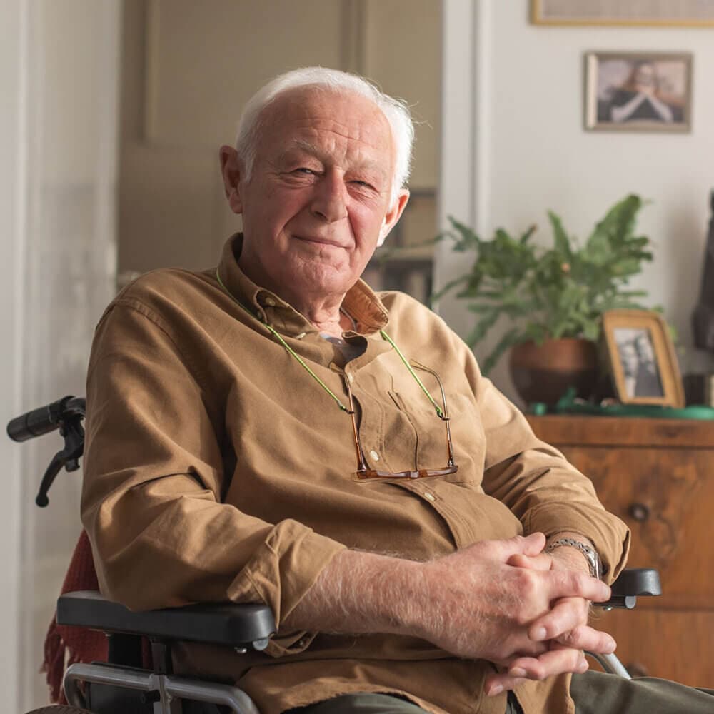 An elderly man with white hair sits in a wheelchair, smiling warmly. A plant and framed photos are in the background. - Home Instead