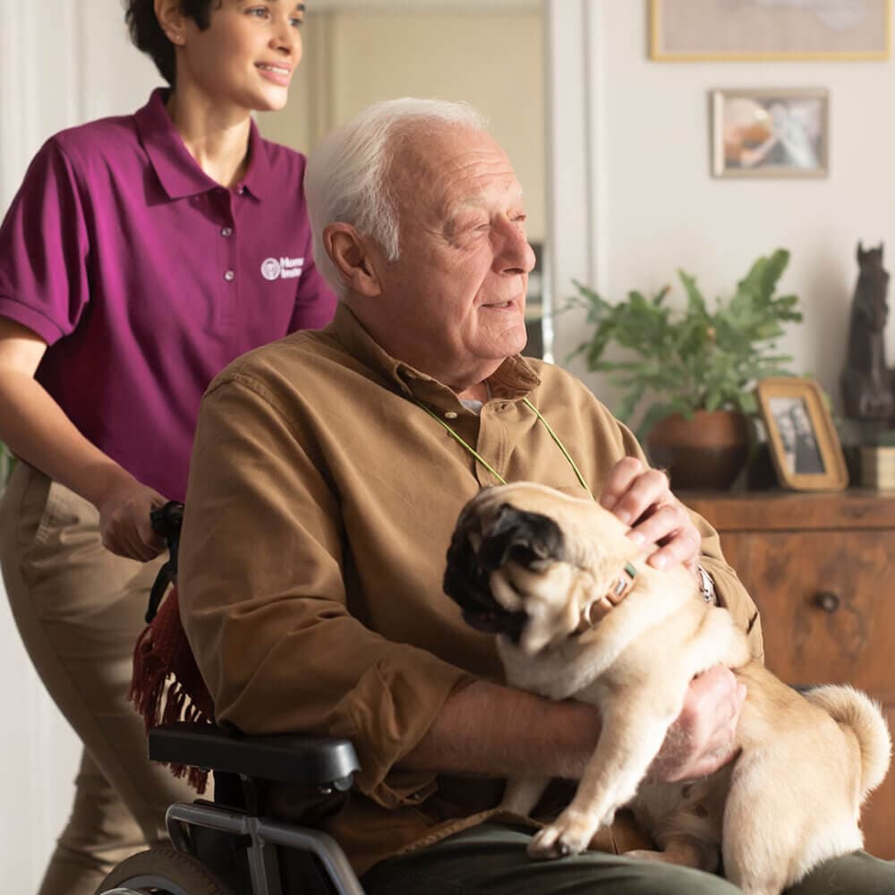 An elderly man in a wheelchair holds a pug, while a caregiver in a purple shirt stands behind him. - Home Instead
