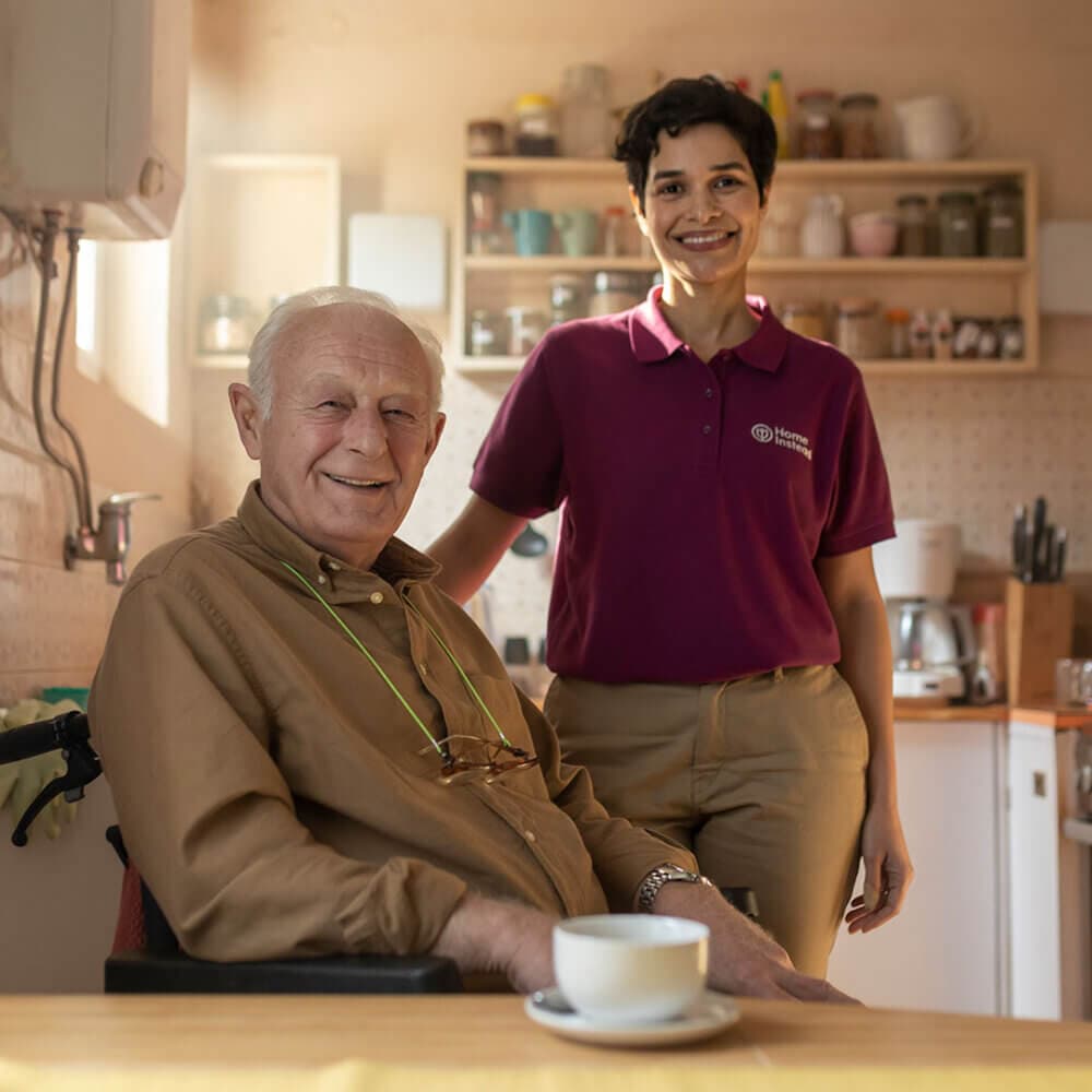 An elderly man in a wheelchair and a caregiver smiling in a kitchen with a cup of coffee on the table. - Home Instead