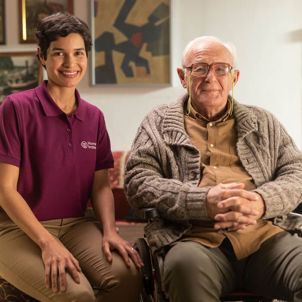 A caregiver in a purple shirt smiles while sitting beside an elderly man in a wheelchair, who is also smiling. - Home Instead