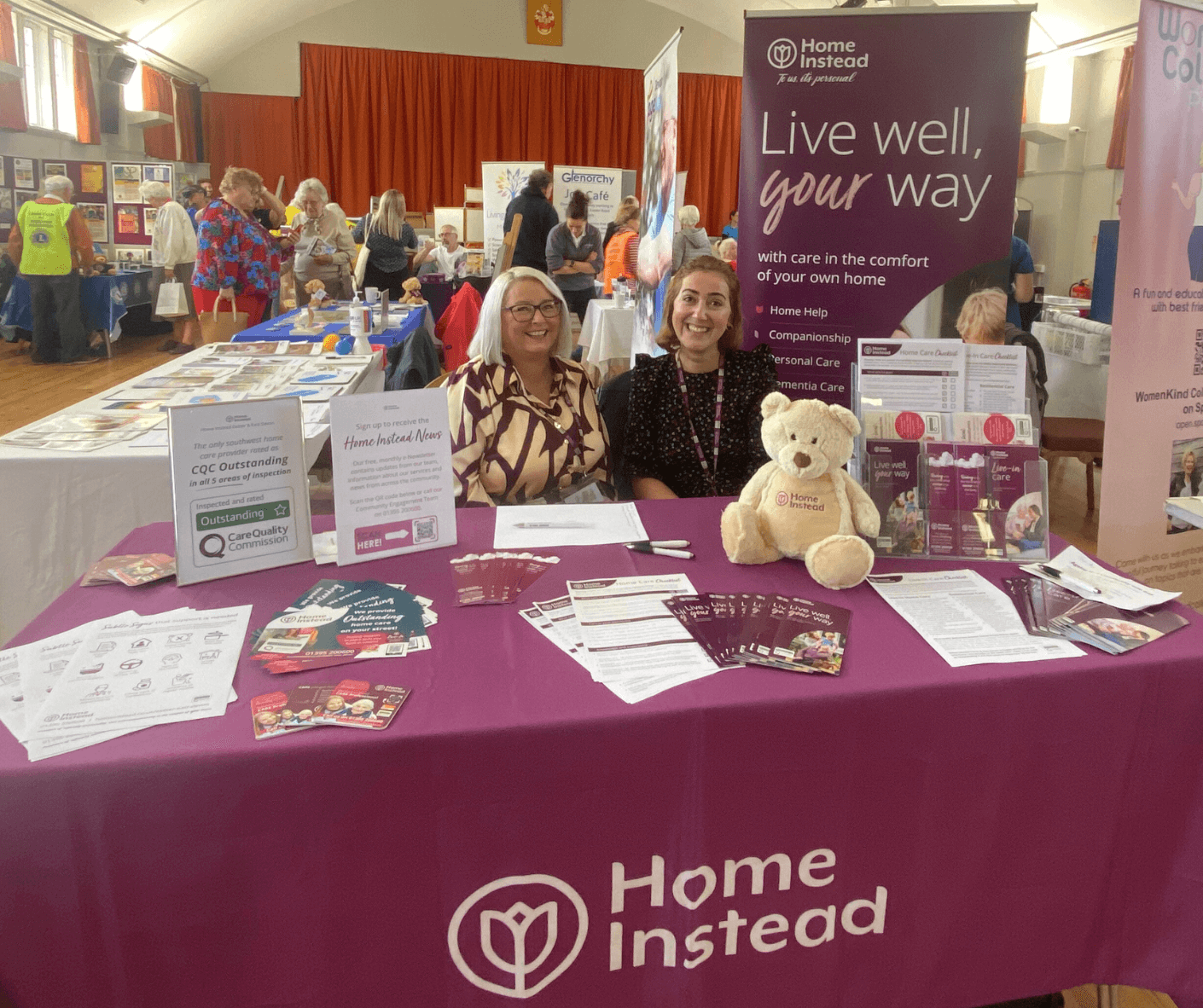 Two women smiling at a Home Instead booth, displaying brochures and a teddy bear, at a caregiving event. - Home Instead