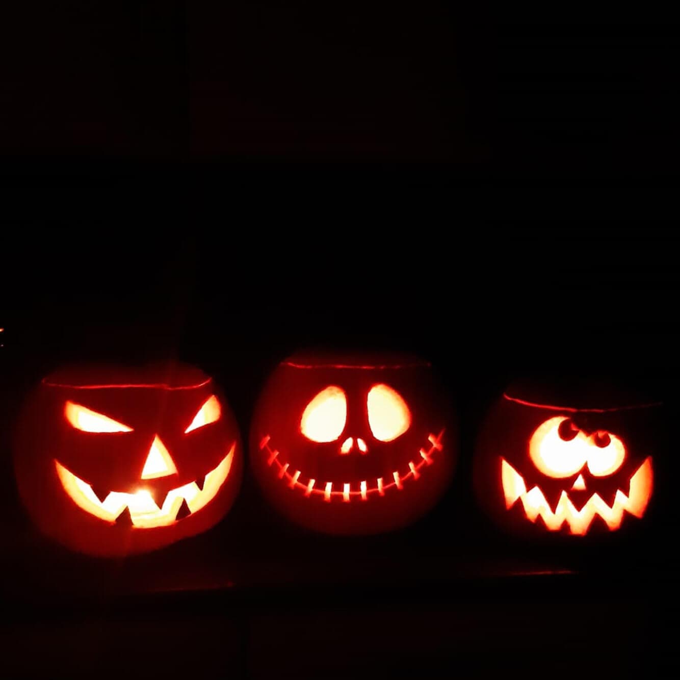 Three glowing jack-o'-lanterns in the dark, each with a different carved face: scary, smiling, and surprised. - Home Instead