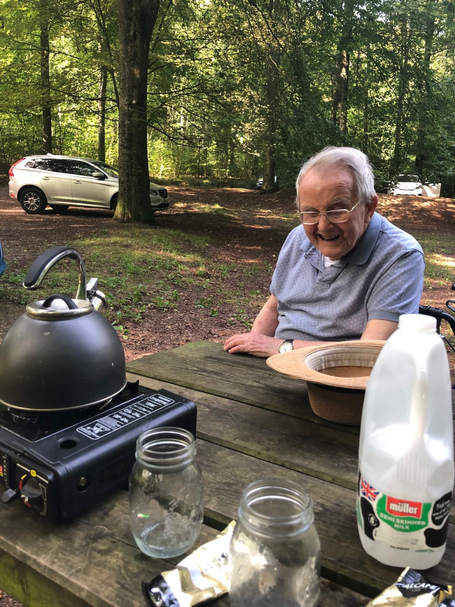Elderly man smiling at a picnic table in a wooded area with a camping stove, kettle, and milk carton. Car in the background. - Home Instead
