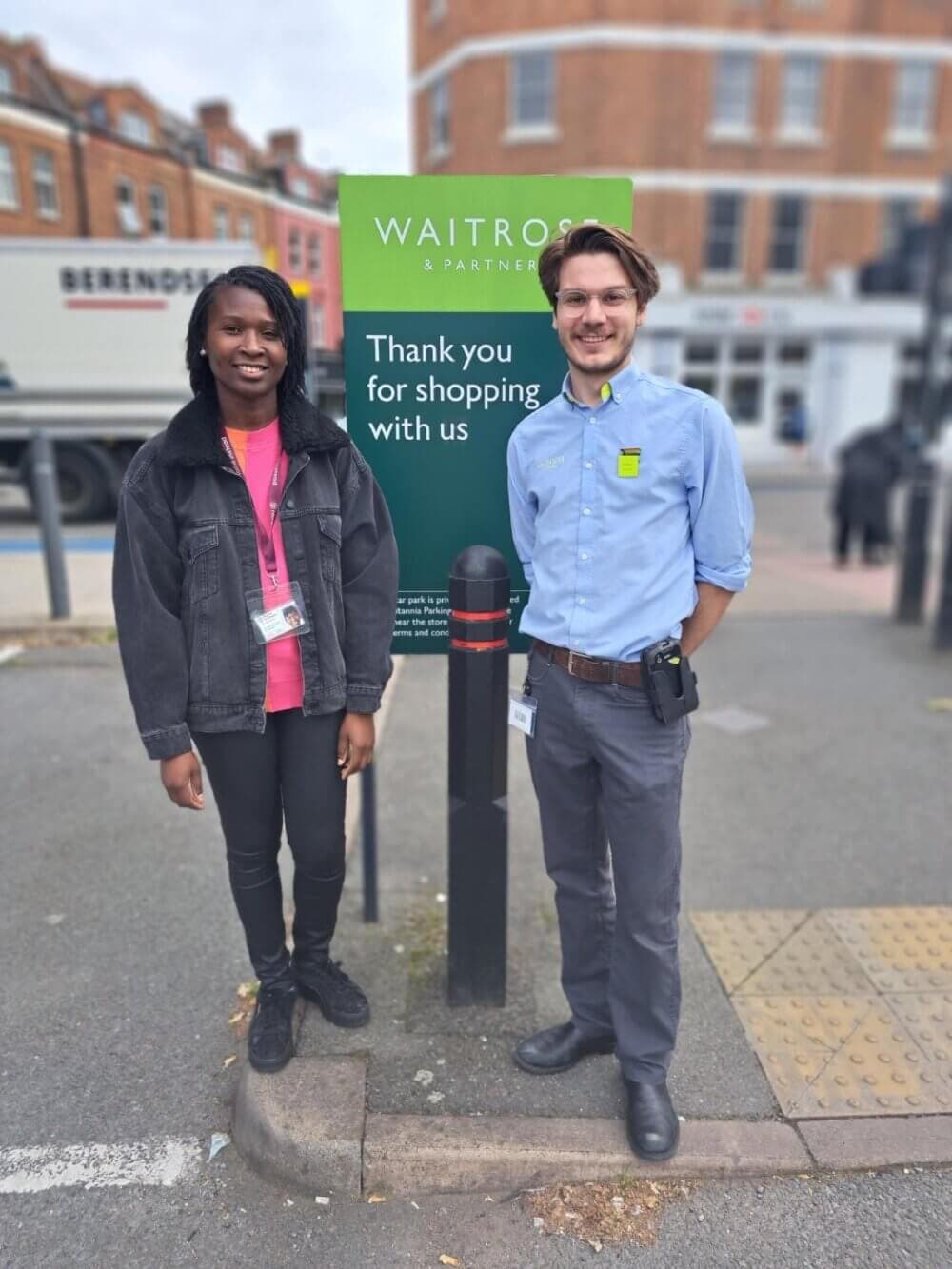 Two store employees standing and smiling outside a Waitrose & Partners store near a "Thank you for shopping with us" sign. - Home Instead