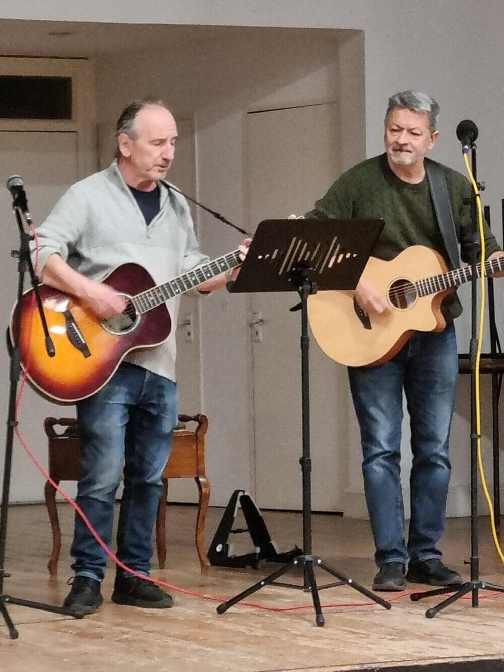 Two men standing and playing guitars on a small indoor stage, with microphones in front of them. - Home Instead