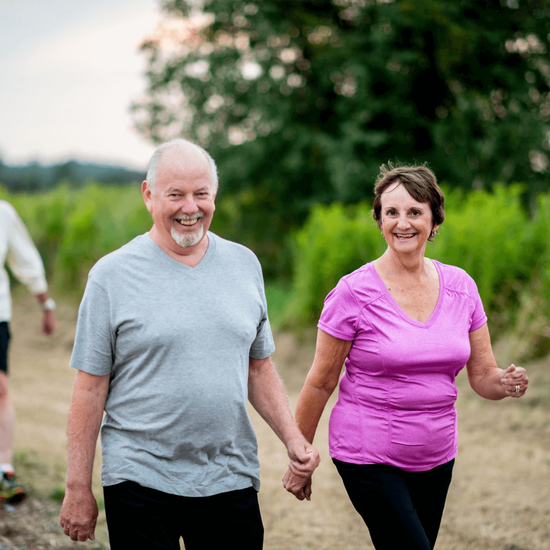 Smiling couple holding hands while walking outdoors in casual attire, with greenery in the background. - Home Instead