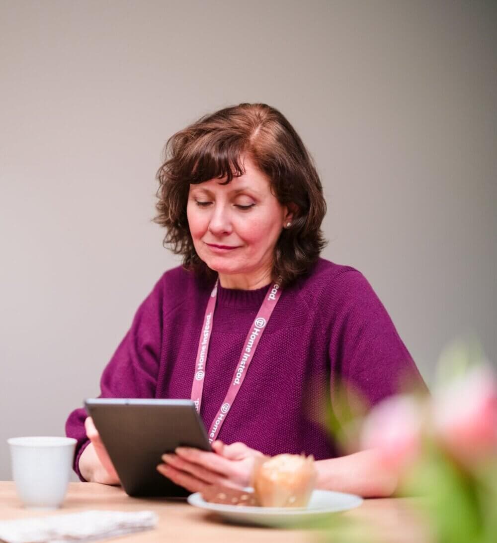 Person with short brown hair wearing a purple sweater, reading a tablet at a table with a cup and pastry. - Home Instead