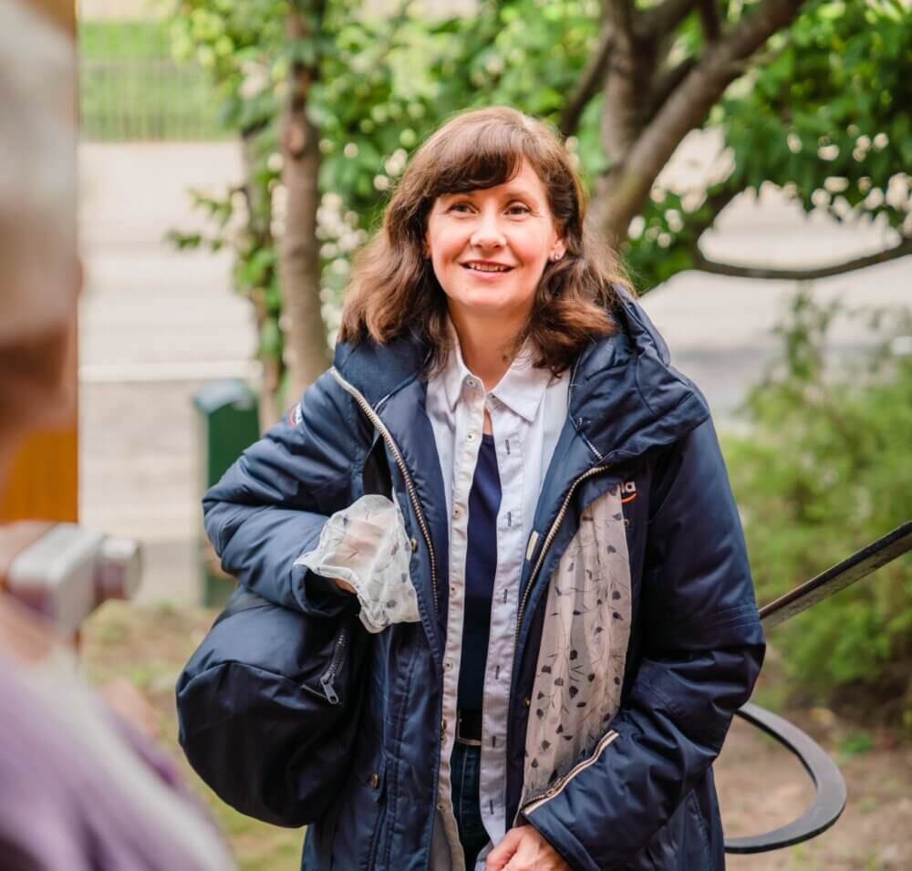 Woman with a smile, wearing a coat and holding belongings, stands outdoors on a pleasant day. - Home Instead