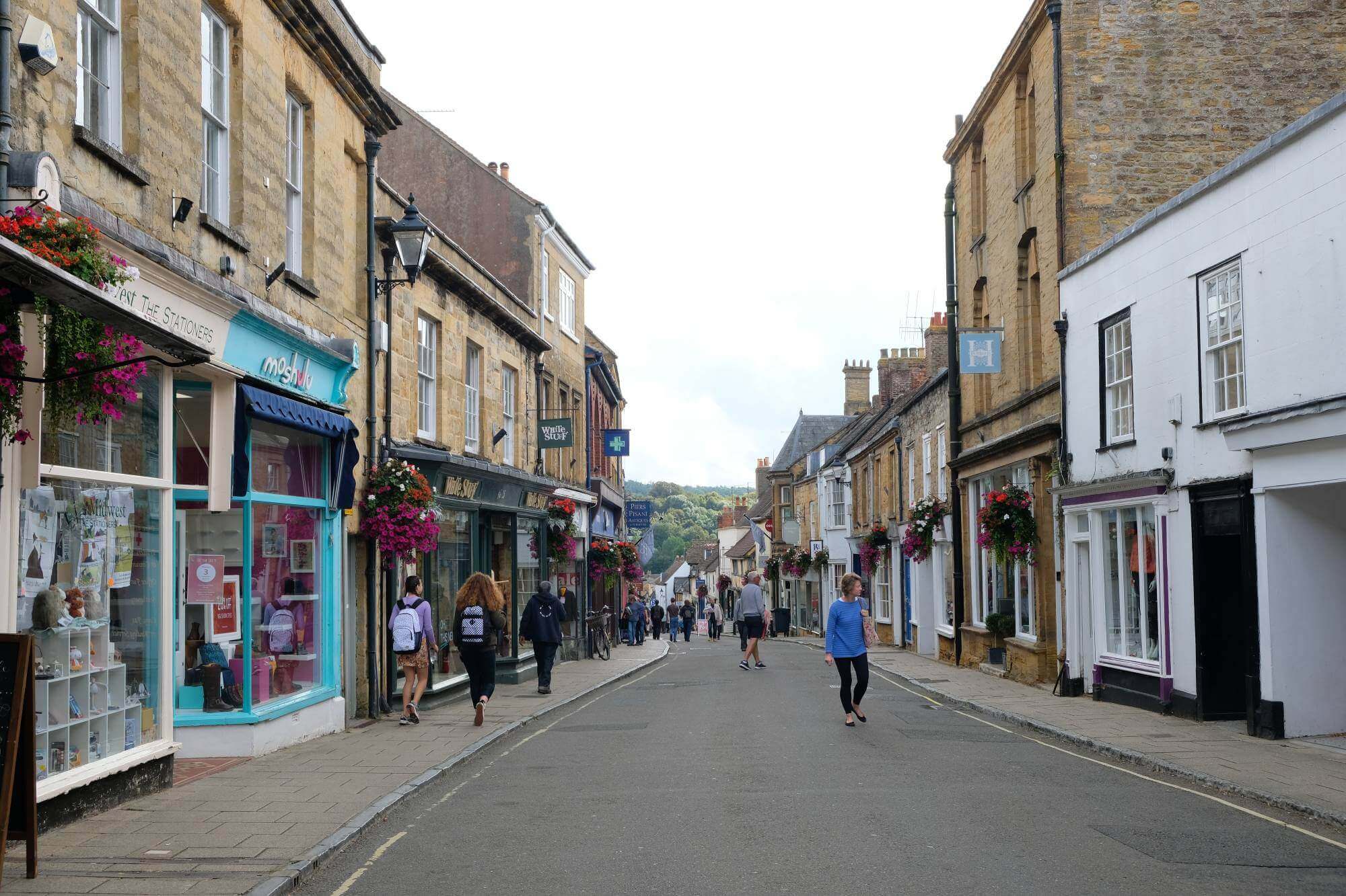 People walk past shops on a quaint, bustling street with historic architecture on a clear day. - Home Instead