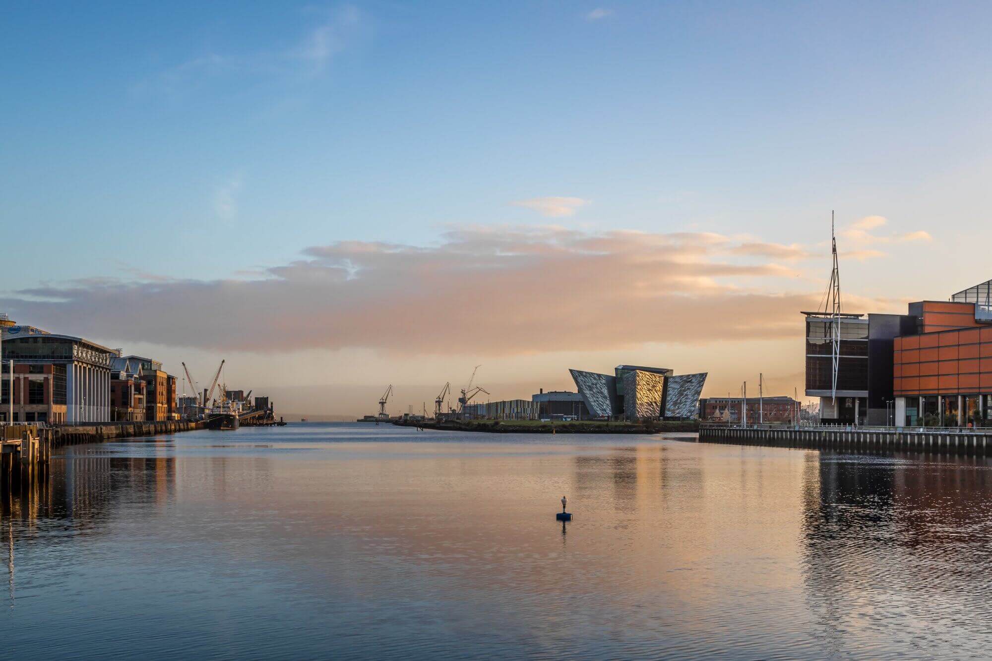 Calm waterfront at sunrise, with modern angular buildings and industrial structures lining the sides. - Home Instead