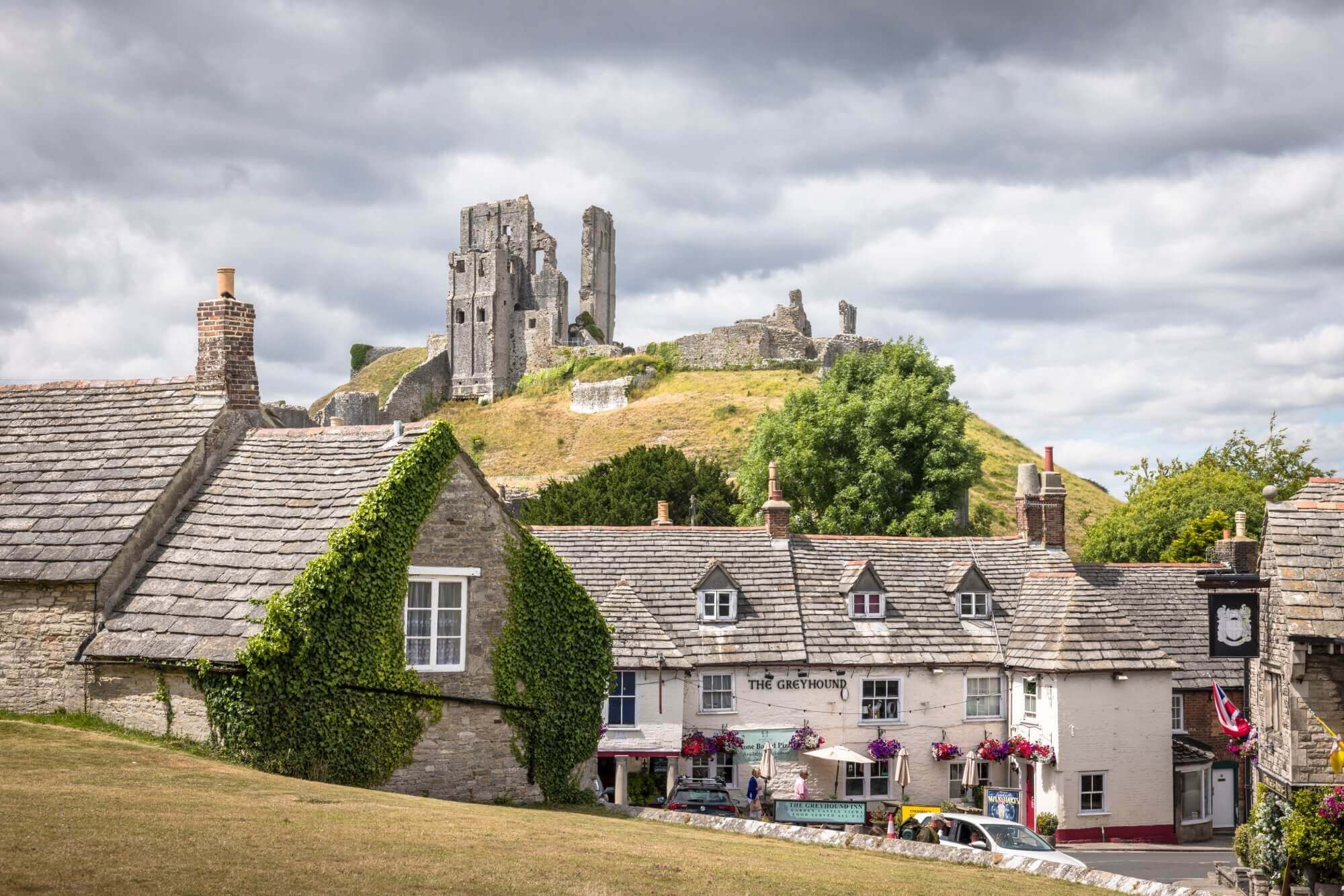A quaint English village with stone houses and a pub, set below a hill with historic castle ruins under a cloudy sky. - Home Instead
