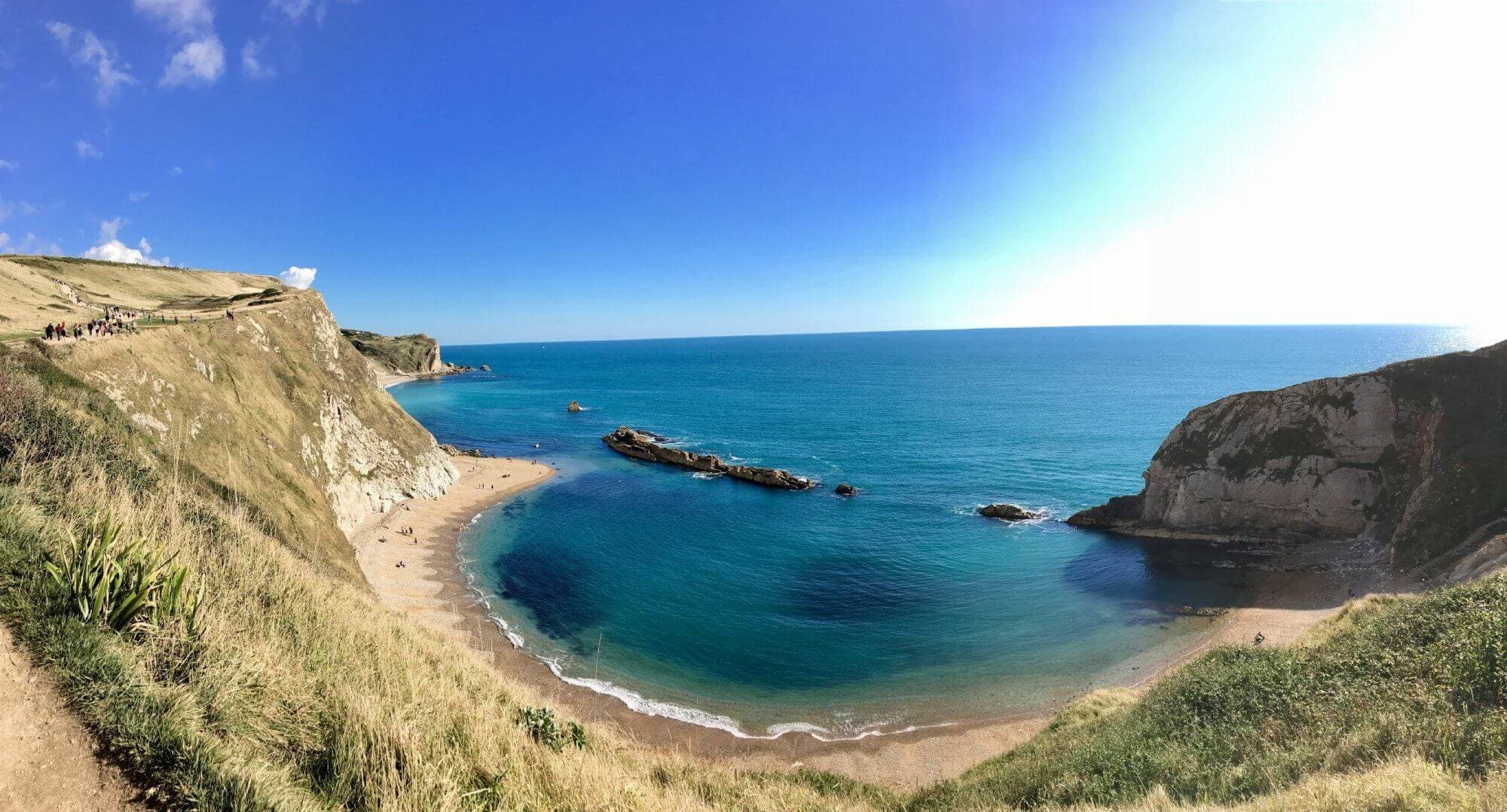 Panoramic view of a serene bay with turquoise waters, rocky cliffs, and a sandy beach under a clear blue sky. - Home Instead